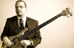A photo of Jason Raso dressed in a suit with a four string bass looking bored.
